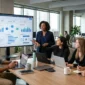 A diverse team of five software developers, project managers, and analysts collaborating around a large monitor displaying a business dashboard in a modern, naturally lit office.