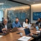 Diverse professionals sitting around a conference table with laptops, viewing global network overlays and data dashboards.