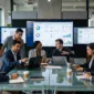A diverse team of software developers and business professionals sitting around a glass conference table collaborating, with large displays showing analytics and enterprise software dashboards in a modern US corporate office.